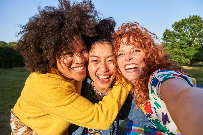 Three young women, one Black, one East Asian and one white, hugging each other and smiling at the camera for a selfie.